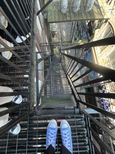 Photograph of a pair of feet in converse sneakers ready to go down a steep iron staircase in a rough city alley.
