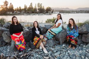 Four professional Indigenous women in ribbon skirts sit on the rocky shores of a lake